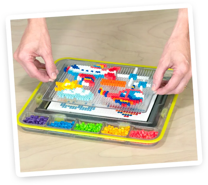 The accessory in use — hands pressing real beads onto the pegboard sitting over the iPad, with beads sorted by color in the tray below