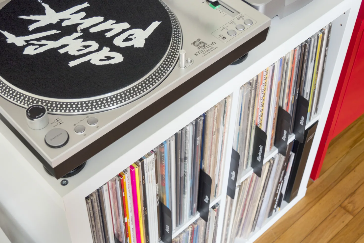 A turntable on top of a white cube shelf of LPs, with black dividers labeled Jazz, Rap, Indie, Electronic, Dance, and Disco sorting the records below