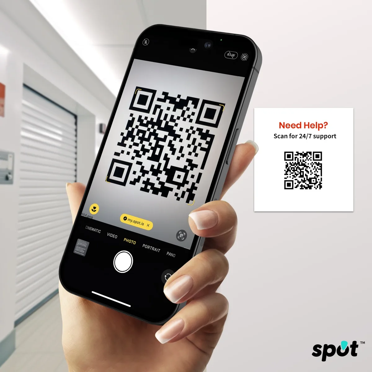 spot in use: a hand holds an iPhone up in a self storage hallway, its camera reading a QR code from a Need Help? Scan for 24/7 support sign on the wall, with a my.spot.is link ready to open