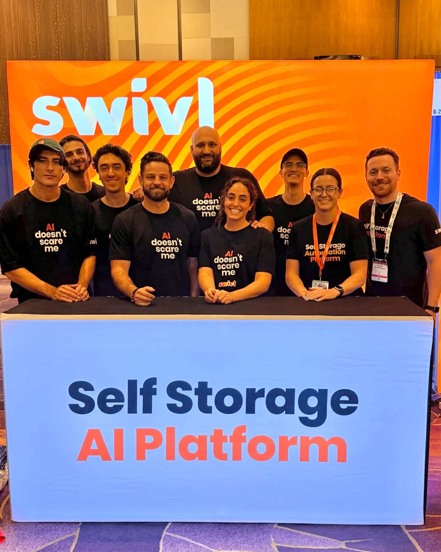 The swivl team at a trade show booth — nine people, most in black &ldquo;AI doesn&rsquo;t scare me&rdquo; T-shirts, lined up behind a table reading Self Storage AI Platform, under an orange swivl banner
